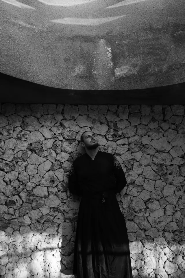 OPPAACHA standing against a rough stone wall under a concrete canopy, black and white