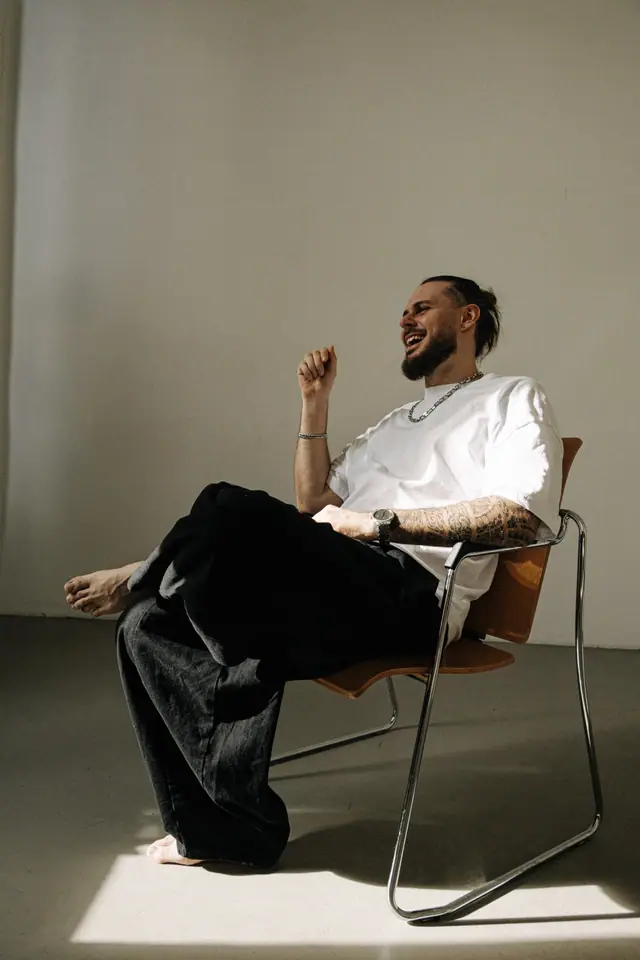 OPPAACHA laughing in a tubular steel chair, barefoot, in daylight